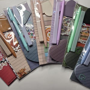May include: A collection of stationery sets in clear plastic packaging, each containing a pen, a notepad, and decorative stickers. The sets feature various colors including tan, light blue, pink, lavender, mint green, and dark blue, with heart-shaped pads.