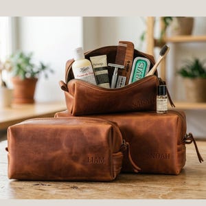 Puede incluir: Tres neceseres de cuero marrón de diferentes tamaños apilados sobre una superficie de madera. La bolsa superior está abierta, revelando productos de aseo. Las bolsas están grabadas con los nombres "SARAH", "LIAM" y "NOAH".