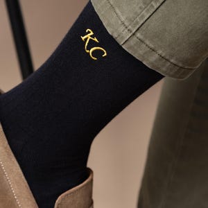 May include: Dark navy ribbed socks featuring the gold embroidered initials "KC". The socks are worn with olive green trousers and a brown shoe, visible on a person's leg.