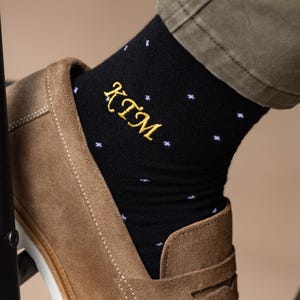 May include: Black socks with white star patterns and gold embroidered initials "KTM". The socks are worn with brown suede loafers and olive-green trousers. A stylish accessory.