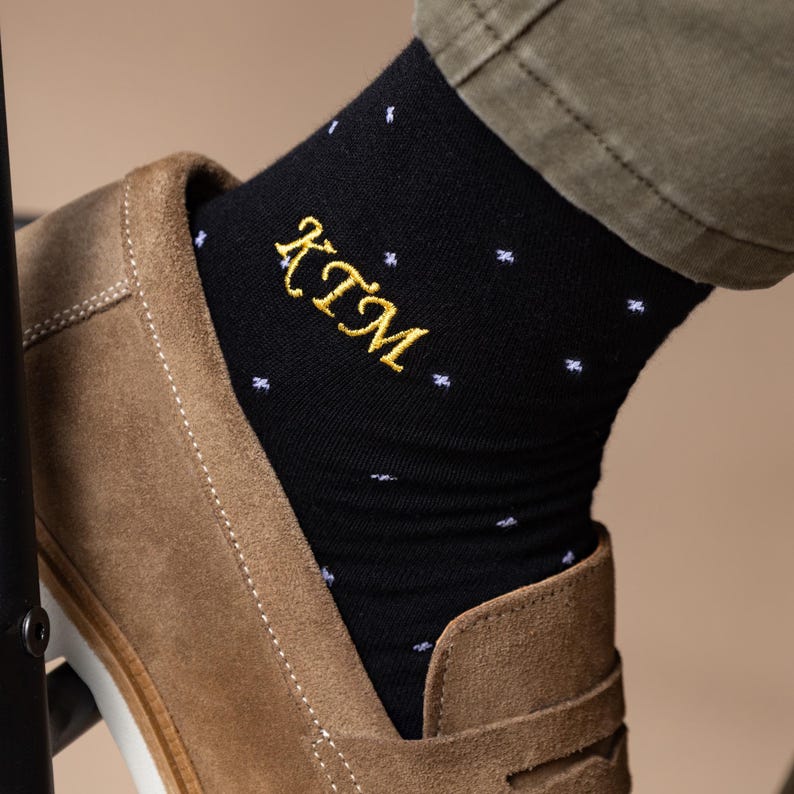 May include: Black socks with white star patterns and gold embroidered initials "KTM". The socks are worn with brown suede loafers and olive-green trousers. A stylish accessory.