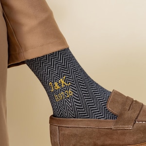 May include: Navy blue and grey patterned socks with gold embroidered initials "J&K" and the date "11.26". The socks are paired with brown suede loafers.