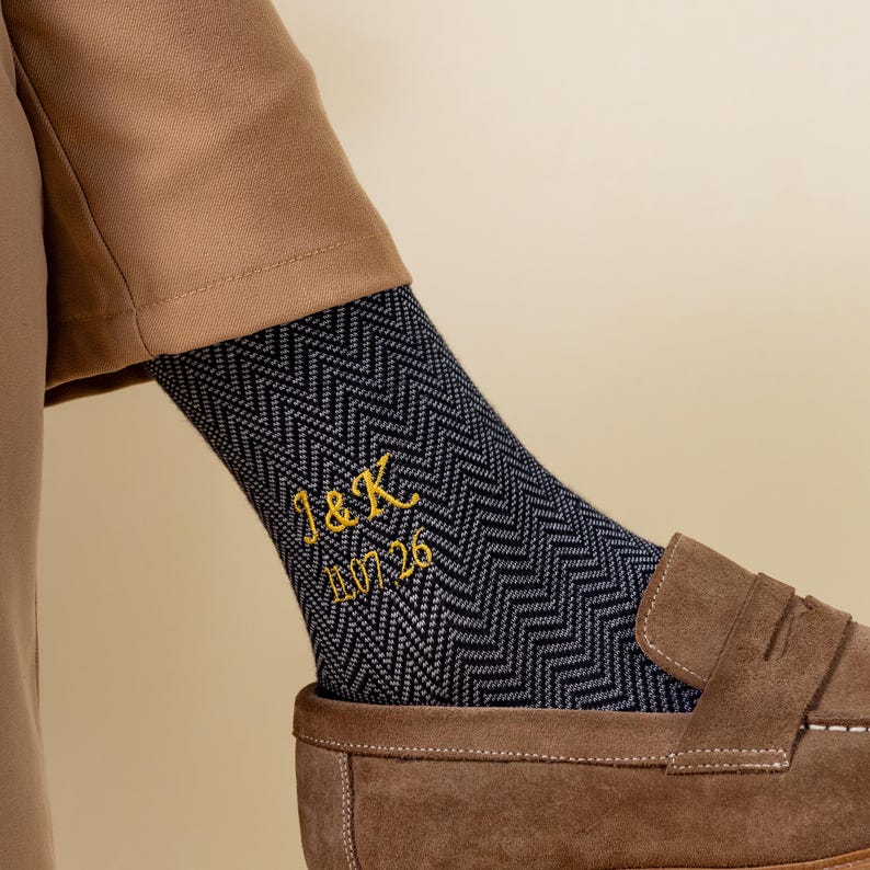 May include: Navy blue and grey patterned socks with gold embroidered initials "J&K" and the date "11.26". The socks are paired with brown suede loafers.