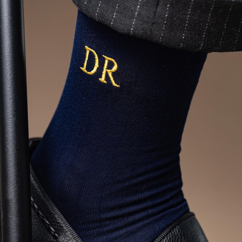 May include: Navy blue socks with gold embroidered initials "DR". The socks are made of a ribbed knit fabric and are worn with black leather shoes and a grey pinstripe suit.