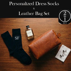 May include: A gift set featuring personalized dress socks and a leather bag. The image displays black socks with "SF" embroidery, a brown leather bag with "SRF" embossed, a small bottle, and cufflinks on a white cloth. The text reads "Personalized Dress Socks & Leather Bag Set".
