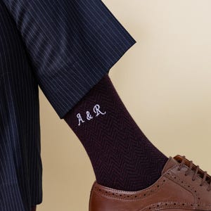 May include: Close-up of burgundy herringbone socks embroidered with the initials "A & R" in white. The socks are worn with brown leather shoes and a navy pinstripe suit. The image focuses on the lower leg and foot.