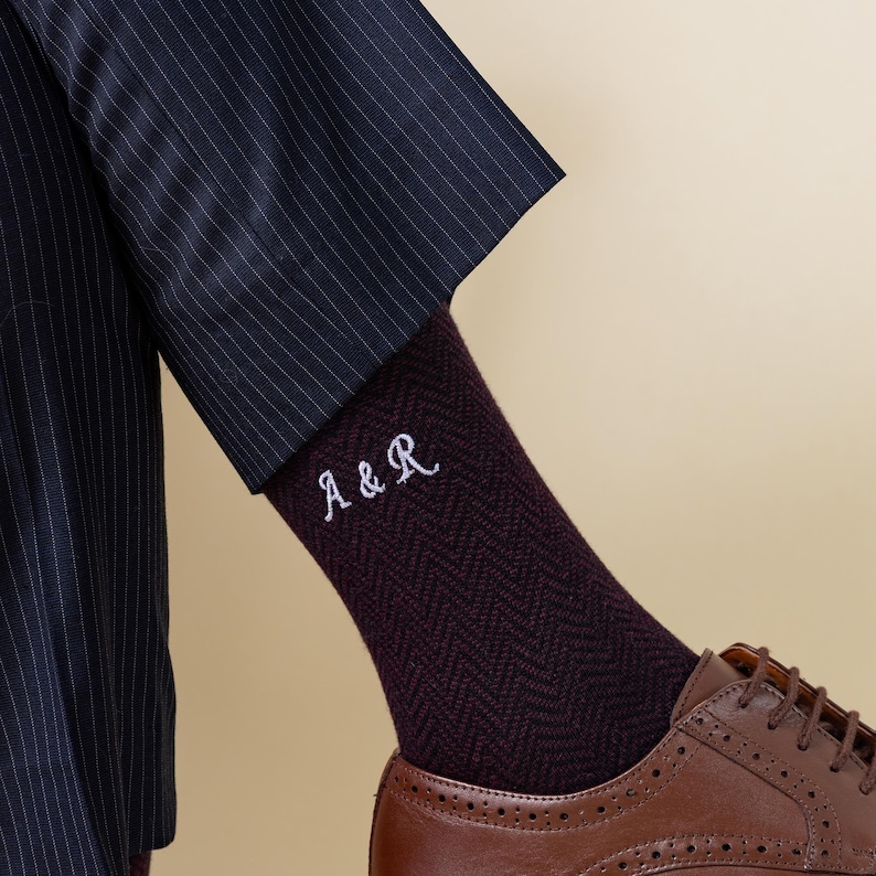 May include: Close-up of burgundy herringbone socks embroidered with the initials "A & R" in white. The socks are worn with brown leather shoes and a navy pinstripe suit. The image focuses on the lower leg and foot.
