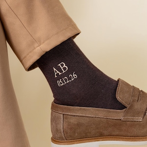 May include: A close-up shot of a brown sock embroidered with the initials "AB" and the date "05.12.26" in white. The sock is worn with a brown loafer and tan trousers. The background is a neutral beige.