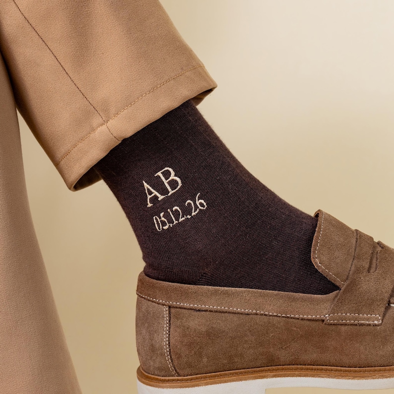 May include: A close-up shot of a brown sock embroidered with the initials "AB" and the date "05.12.26" in white. The sock is worn with a brown loafer and tan trousers. The background is a neutral beige.