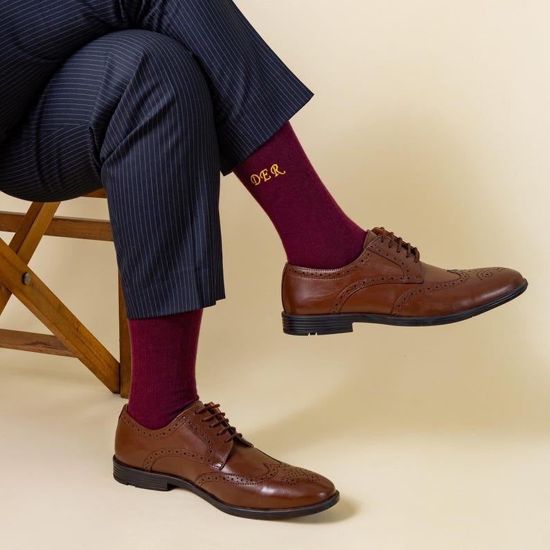 May include: A person seated, wearing navy pinstripe trousers, burgundy socks with "DER" embroidered in gold, and brown leather brogue shoes. The shoes have a classic wingtip design and are laced up. The background is a solid, neutral colour.
