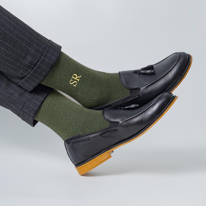 May include: Black leather loafers with tassel detailing, worn with green herringbone socks embroidered with "SR" in gold. The shoes have a light brown sole. The socks are visible above the shoes, set against a neutral background.