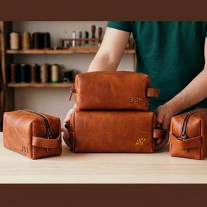 May include: Four rectangular brown leather toiletry bags with zippered closures and side handles. Each bag features a unique embossed monogram: SRF, AK, and JM. The bags have a smooth, matte finish and are arranged on a light-colored surface.