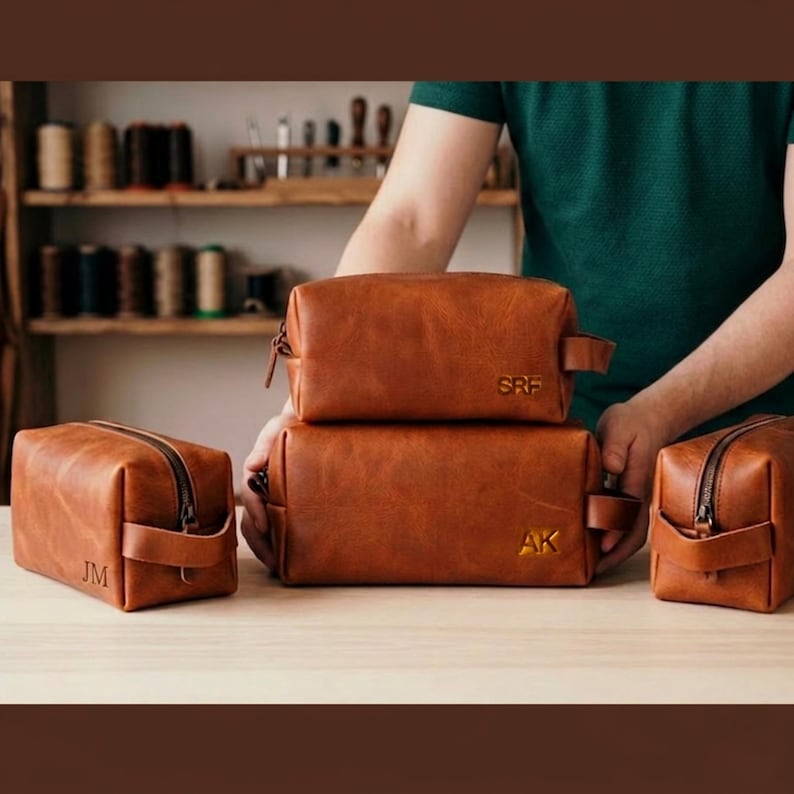 May include: Four brown leather wash bags of different sizes are shown. Each bag has a zip closure and a handle. Two bags are stacked, and each bag has initials embossed on the front. The bags are on a wooden surface.