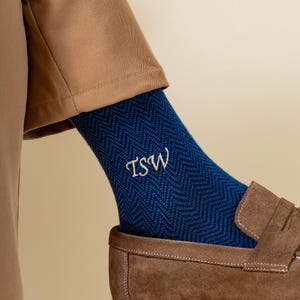 Puede incluir: Calcetines azules con estampado de espiga y las iniciales "TSW" bordadas en blanco. Los calcetines se llevan con mocasines de ante marrón y una camisa beige. Un accesorio elegante.