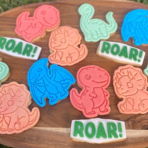 Peut inclure: Assortiment de biscuits en forme de dinosaures aux couleurs vives, rose, orange, bleu et vert, disposés sur un plateau en bois. Certains biscuits affichent le mot "ROAR!".