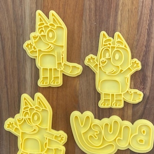May include: A set of four yellow plastic cookie cutters featuring three distinct cartoon dog characters in various poses and one cookie cutter shaped like the word Bluey in a stylized, rounded, and bold bubble font with a connected base.