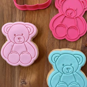 May include: Pink and blue teddy bear-shaped cookies and cookie cutters on a wooden surface. The cookies are decorated with a pink and blue frosting, and the cookie cutters are pink. The cookies are in the shape of a teddy bear.