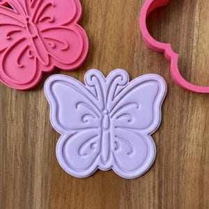 May include: Three butterfly-shaped cookie cutters in shades of pink and purple. The cutters are made of plastic and have detailed designs. The butterfly cookie cutters are on a wooden surface.