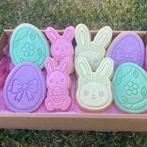 Puede incluir: Una caja de galletas de Pascua con forma de conejos y huevos. Las galletas están decoradas con colores pastel, incluyendo rosa, verde y morado. Las galletas de conejito tienen caras detalladas y detalles florales.
