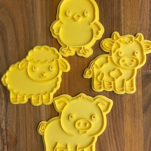 May include: Four yellow plastic cookie cutters shaped like farm animals: a chick, a sheep, a cow, and a pig. The cutters are on a wooden surface. The chick has a beak and feet. The sheep has a fluffy body. The cow has spots. The pig has a curly tail.