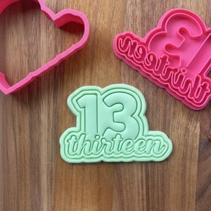 May include: A selection of cookie cutters in pink and green, arranged on a wooden surface. The cutters include a heart shape, the number "13", and the word "thirteen". The "13" and "thirteen" cutters are in a single design.