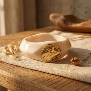 Può includere: Un braccialetto spesso color crema con un accento circolare strutturato color oro. Il braccialetto è realizzato in un materiale liscio e lucido e poggia su una superficie di lino dai toni neutri.