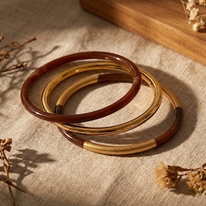 以下が含まれることがあります： 3つの重ねられたバングル。1つは濃い茶色で、他の2つは金色です。茶色のバングルには金色のアクセントがあります。バングルはニュートラルカラーの表面に置かれています。