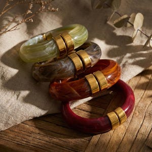 May include: Four resin bangles in green, brown, orange, and red, each with a gold-coloured metal accent. The bangles are displayed on a wooden surface with a neutral-coloured fabric.