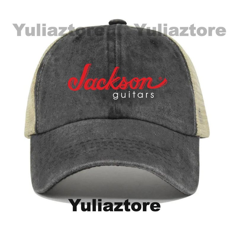 Pu&ograve; includere: Un cappellino da baseball grigio antracite con retro in rete beige e visiera curva. Il cappellino presenta il testo rosso "Jackson" in corsivo sopra la parola "guitars" in bianco. Un classico cappellino trucker.
