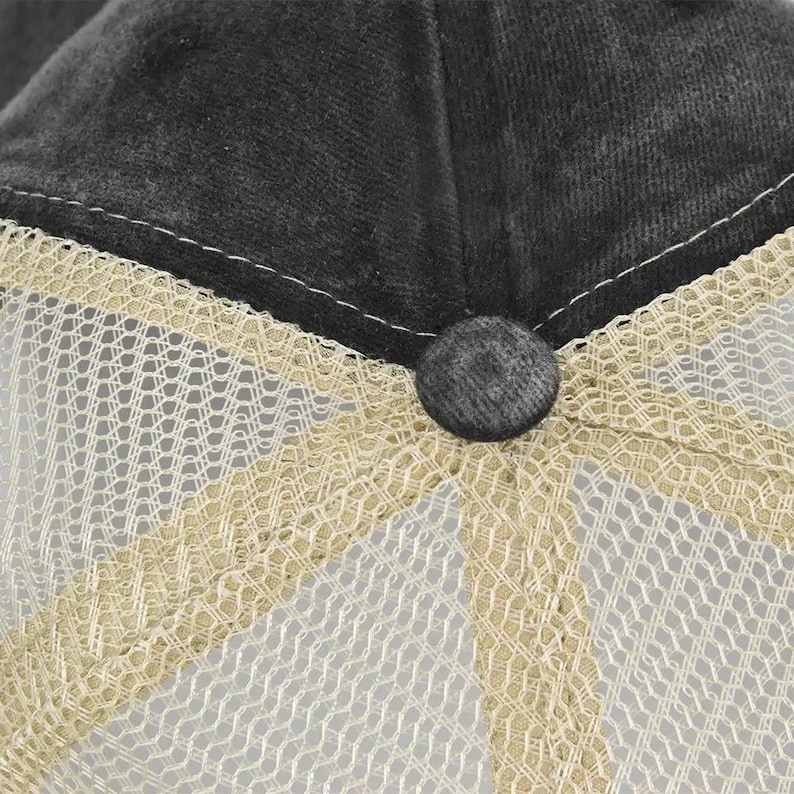 Pu&ograve; includere: Primo piano di un cappellino da camionista grigio antracite. Il cappello presenta una corona in tessuto grigio scuro e un retro in rete beige. Un piccolo bottone rotondo grigio &egrave; centrato sul davanti, collegando la corona alla rete.