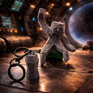 Op de afbeelding: Een grijze, 3D-geprinte figuur met een uitgestrekte arm, staand op een groene basis, naast een bijpassende sleutelhanger. Het beeldje en de sleutelhanger hebben een geometrisch ontwerp. De achtergrond is een vage, futuristische omgeving.