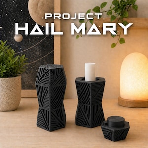 Op de afbeelding: Zwarte geometrische kandelaars met een textuurontwerp. Eén houdt een witte kaars vast. De tekst "PROJECT HAIL MARY" wordt boven de kandelaars weergegeven. Een kleine potplant en een decoratieve lamp staan ook in de afbeelding.