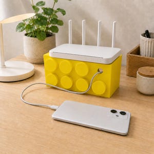 Könnte beinhalten: Eine leuchtend gelbe, rechteckige Aufbewahrungsbox im Design eines Bausteins mit acht kreisförmigen Noppen auf der Vorderseite. Ein weißer WLAN-Router mit vier vertikalen Antennen steht darauf und ein graues Ladekabel verbindet ein silbernes Smartphone.