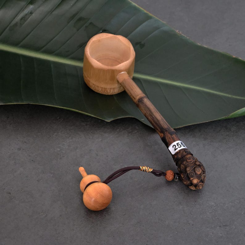 May include: A bamboo water ladle with a cylindrical cup and a dark, textured handle featuring a white sticker with the number 255. A small, light-colored wooden gourd-shaped charm is attached to the end of the handle by a dark brown cord.