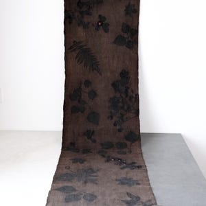 May include: A long, rectangular brown textile with a botanical print of leaves and flowers in black. The textile is displayed vertically, with a raw edge on one side. The background is white and gray.