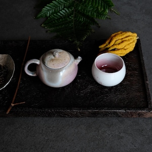 Puede incluir: Una tetera de cerámica con esmalte suave rosa y beige, una taza pequeña a juego con interior rosa intenso, una bandeja de madera marrón oscura, un fruto de mano de Buda amarillo seco, una hoja de helecho verde y un cuenco con té.