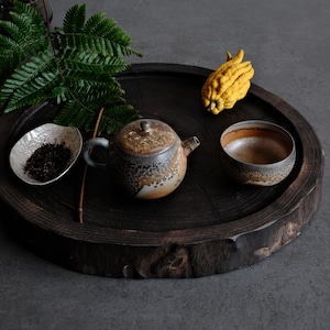 May include: A round dark wood tray holds a ceramic teapot with a mottled brown and cream glaze, a matching tea bowl, a small irregular white dish containing loose tea leaves, a green fern frond, and a yellow Buddha's hand citron fruit.