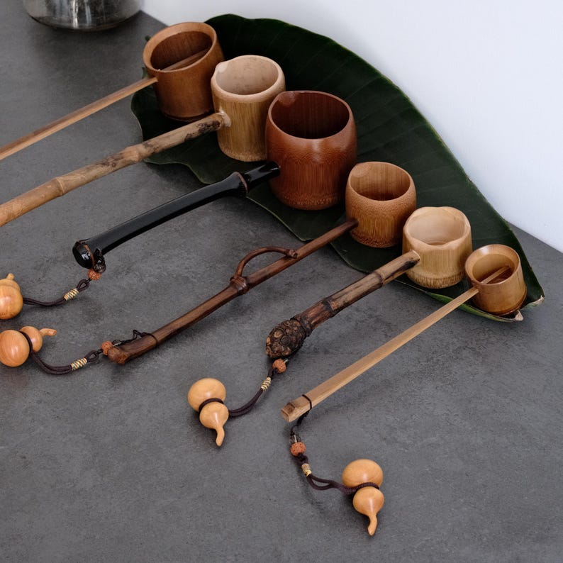 May include: A collection of five bamboo water ladles with varying handle textures and wood tones. Each ladle features a cylindrical cup and a long handle, with some handles finished in black or dark brown, and small gourd-shaped charms attached.