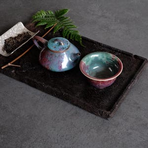 Op de afbeelding: Een keramische theepot en bijpassende theekom met een glanzend, gevlekt glazuur in tinten blauwgroen, blauw en magenta. De set staat op een donker, getextureerd rechthoekig dienblad naast een klein wit schaaltje met losse theeblaadjes en een varenblad.
