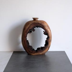Puede incluir: Un jarrón de madera hueco de forma ovalada con un acabado de veta de madera natural en color marrón oscuro. El centro del jarrón presenta una gran abertura irregular con bordes dentados y rugosos, y la parte superior tiene una pequeña abertura.