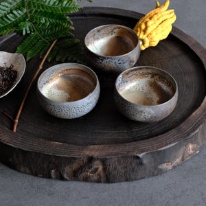Può includere: Tre ciotole in ceramica rotonde con una smaltatura grigia e marrone maculata e una finitura interna liscia. Le ciotole sono disposte su un vassoio circolare in legno scuro insieme a una foglia di felce, un piattino di tè e un cedro giallo.