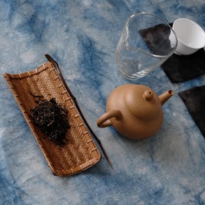 May include: A still life arrangement featuring tea preparation items. A small, round, brown teapot sits near a clear glass pitcher and a white teacup. A woven bamboo tray holds loose tea leaves, with a wooden stick alongside.