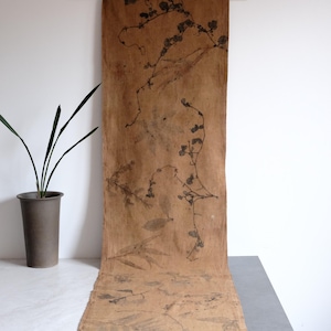 May include: A long, rectangular piece of tan fabric featuring dark brown botanical prints of various leaves and stems scattered across its surface. The fabric has a raw, unfinished edge and displays a natural, earthy colour palette throughout.