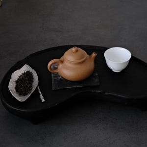 Könnte beinhalten: Eine braune Keramik-Teekanne mit passendem Deckel steht auf einem kleinen quadratischen Untersetzer. Eine weiße Teetasse und eine silberne, blattförmige Schale mit Teeblättern befinden sich auf einem schwarzen Holztablett. Teeservice.