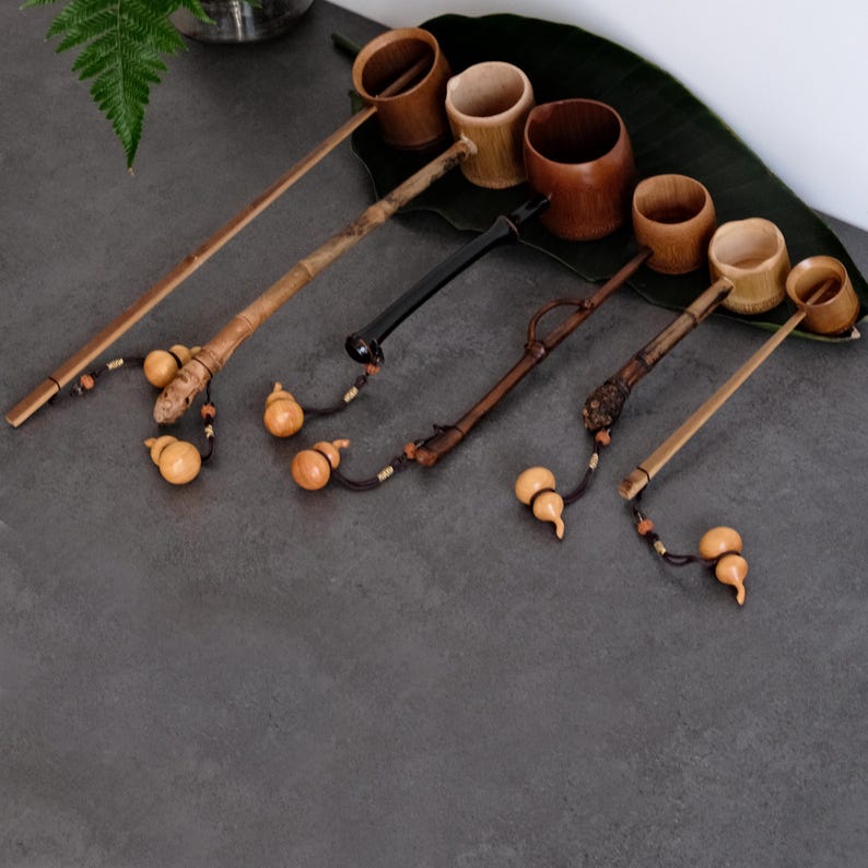 May include: A collection of five wooden ladles with long, slender handles of varying lengths and textures. Each ladle features a small, hollowed-out wooden cup at one end and a decorative, light-colored gourd-shaped charm attached by a dark cord.