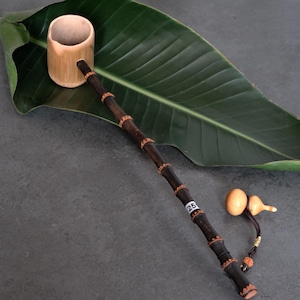 May include: A bamboo ladle featuring a light tan cylindrical cup and a long, dark brown bamboo handle with segmented nodes. A small white sticker with the number 25 is attached to the handle. A small tan gourd charm hangs from the end.