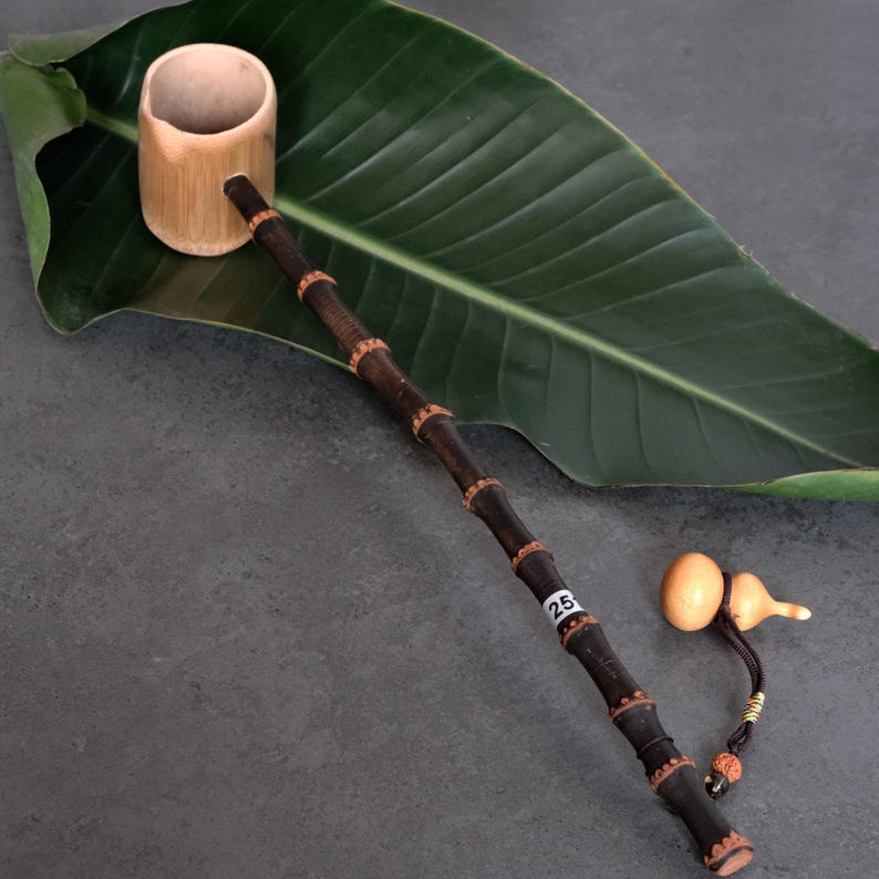 May include: A bamboo ladle featuring a light tan cylindrical cup and a long, dark brown bamboo handle with segmented nodes. A small white sticker with the number 25 is attached to the handle. A small tan gourd charm hangs from the end.