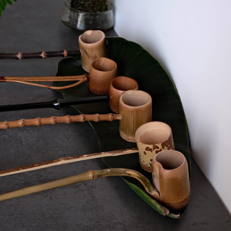 May include: A collection of six bamboo ladles with long, slender handles and cylindrical cups of varying textures and shades of brown, ranging from light tan to dark, polished black, arranged in a fan-like pattern on a large, dark green leaf.