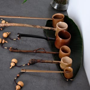 May include: A collection of five bamboo water ladles with long handles, each featuring a unique natural texture and color ranging from light tan to dark brown. Each ladle is accented with a small decorative gourd tied with a dark cord.