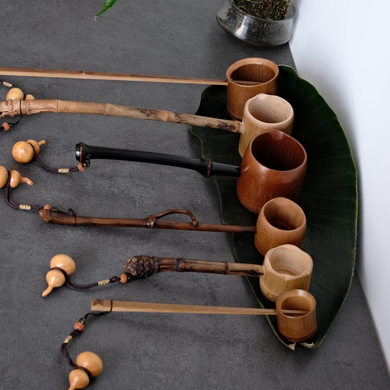 May include: A collection of five bamboo water ladles with long handles, each featuring a unique natural texture and color ranging from light tan to dark brown. Each ladle is accented with a small decorative gourd tied with a dark cord.